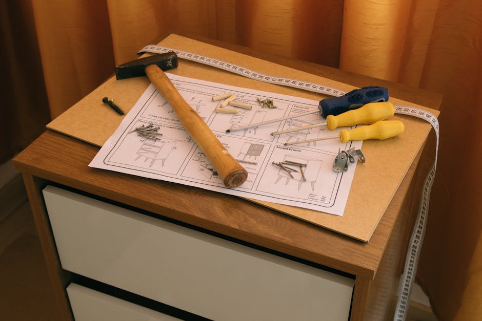 A man with dark hair and a beard, wearing a red sweater, is lifting and carrying a small wooden table in an interior room of a home during a moving or furniture disassembly process. The room features 